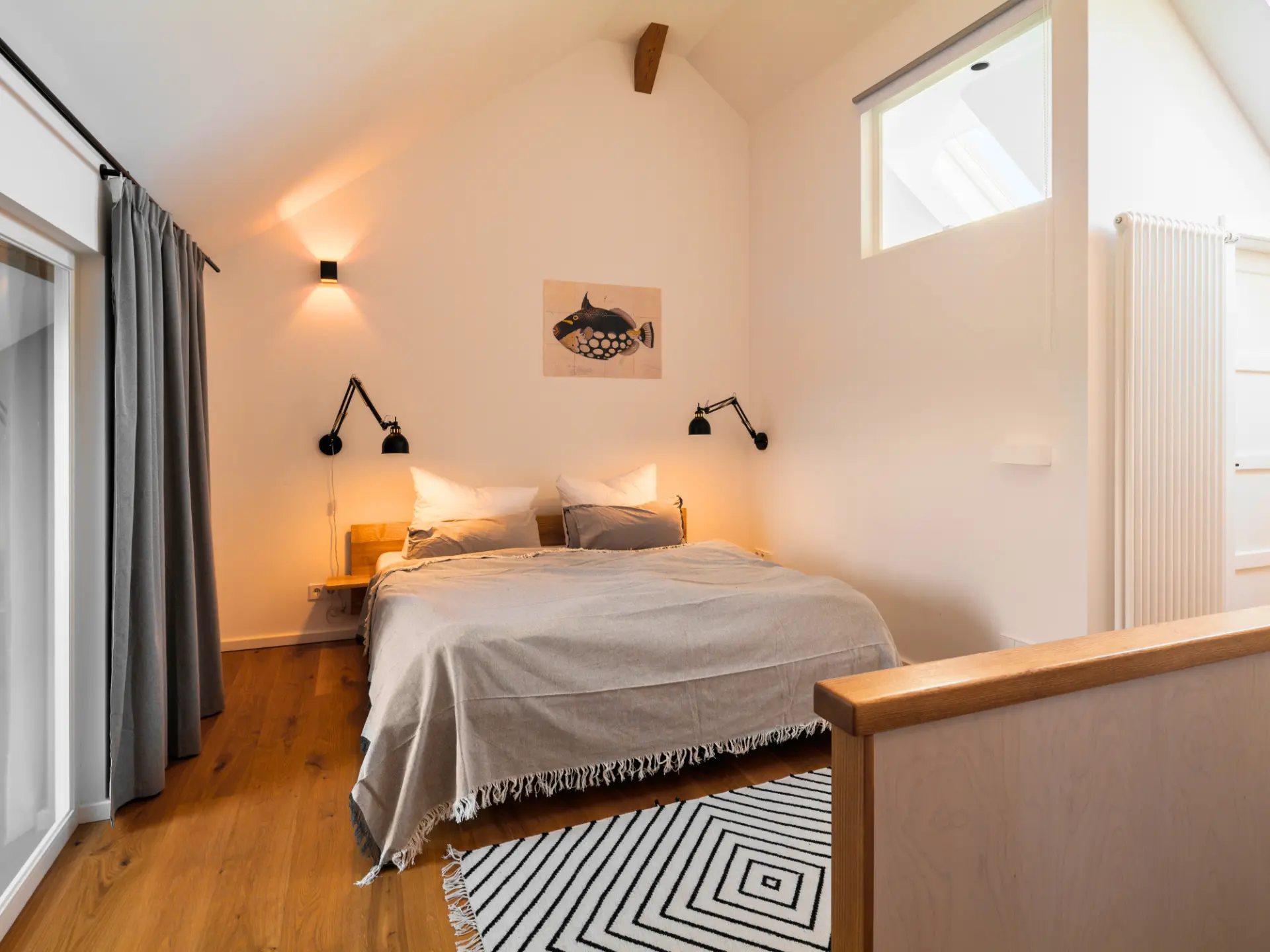 Zweites Schlafzimmer im Obergeschoss mit Doppelbett unter der hohen Giebeldecke, schwarz-weiß gemustertem Teppich und bodentiefem Fenster