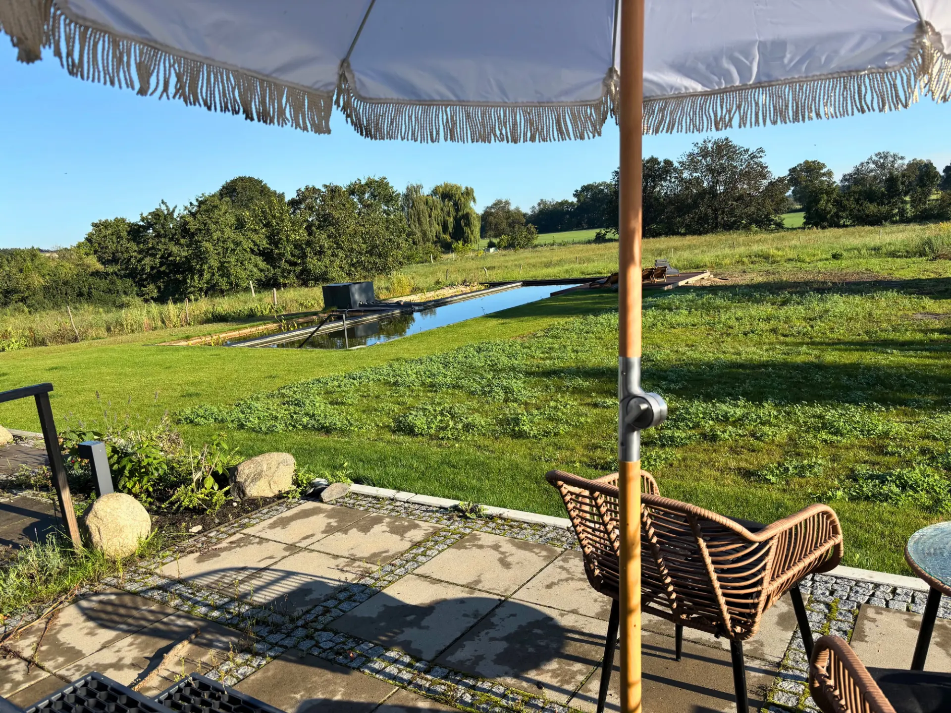 Terrasse am Naturpool: Sonnenschirm, Rasen und Schwimmteich vor den Feldern