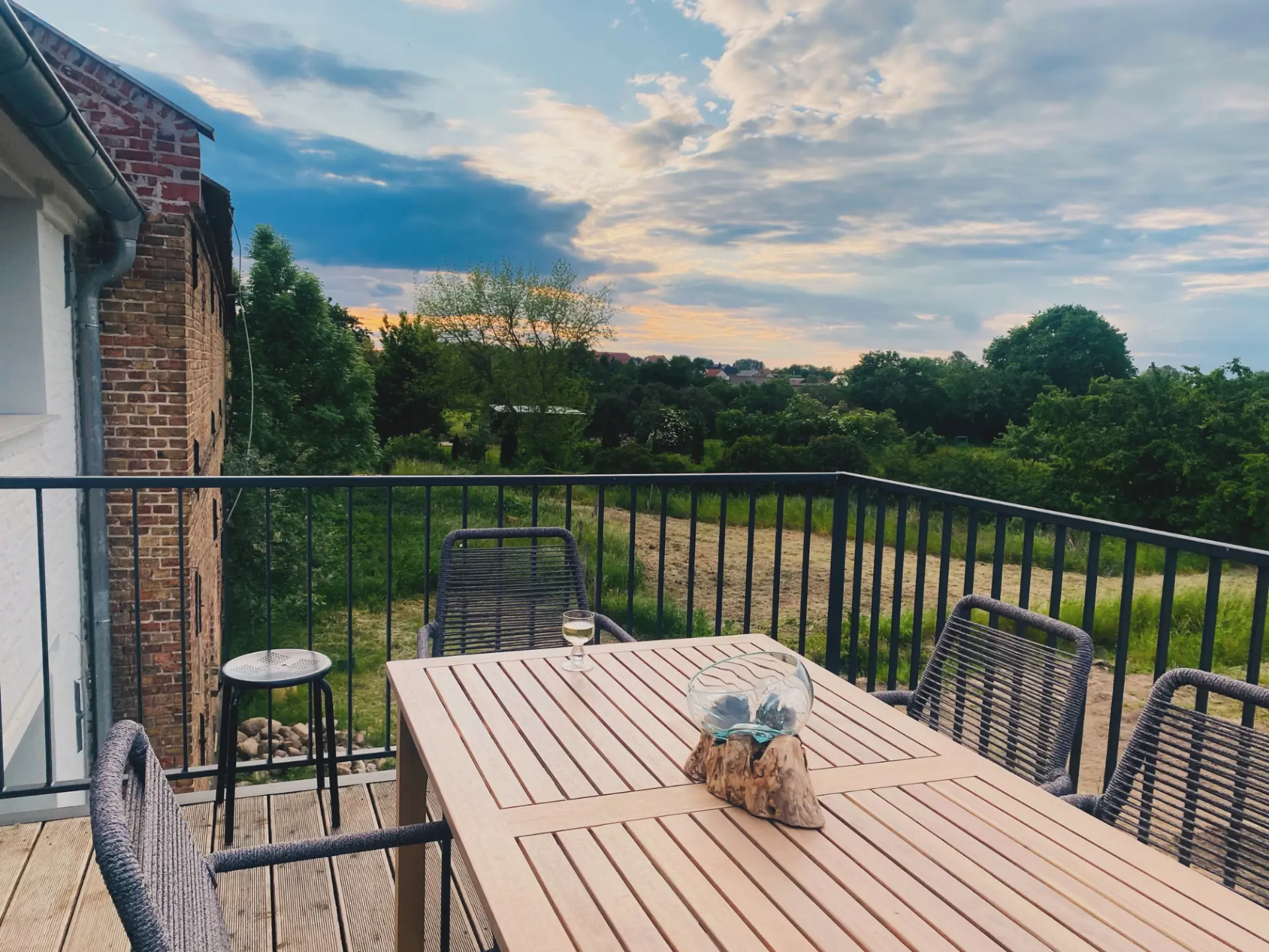 Holzterrasse mit langem Esstisch und geflochtenen Stühlen, Blick über Wiesen und Felder bis zum Abendhimmel